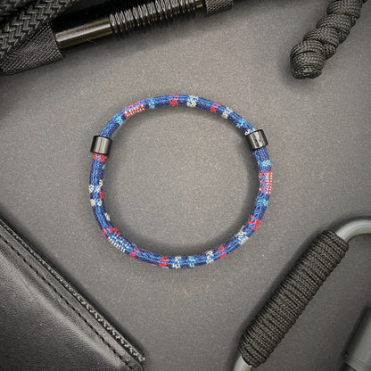 Dark Blue and red Boho rope bracelet with black sliding clasps, displayed on a dark grey surface with EDC gear in the background.