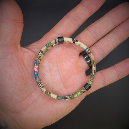 Khaki Boho rope bracelet with black sliding clasps, displayed in an open palmed hand over a dark grey surface.