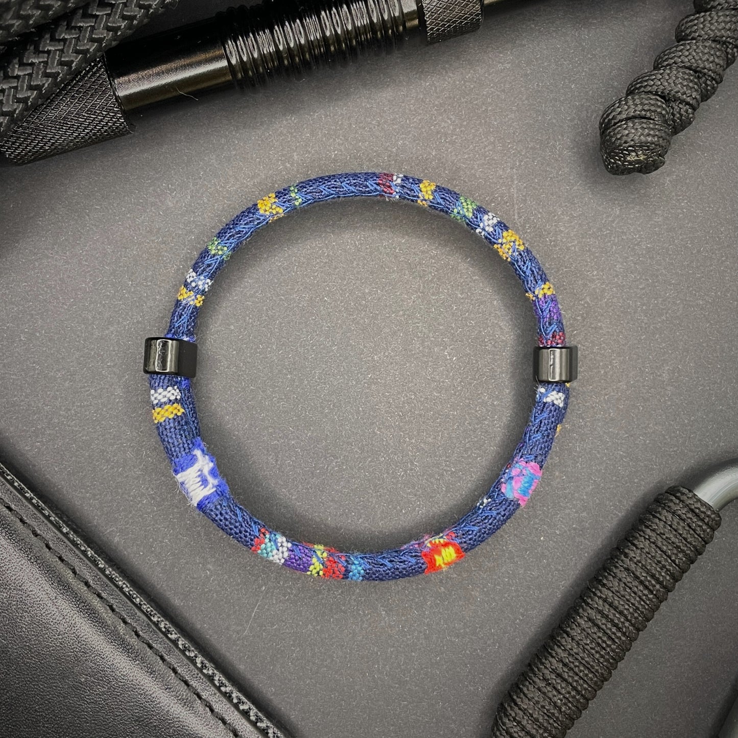 Dark Blue Boho rope bracelet with black sliding clasps, displayed on a dark grey surface with EDC gear in the background.