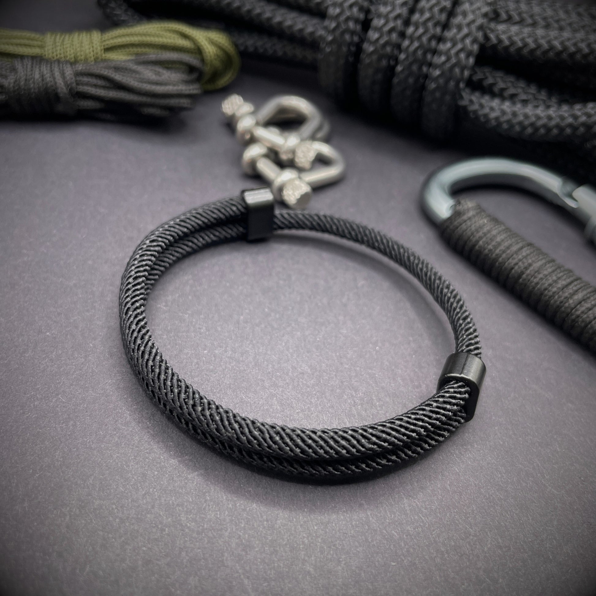 Black Milan rope bracelet with black sliding clasps, displayed on a dark grey surface with EDC gear in the background.