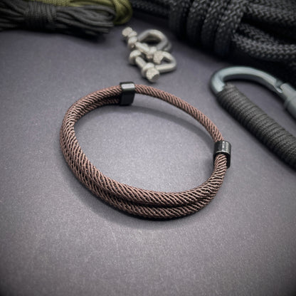 Coffee Milan rope bracelet with black sliding clasps, displayed on a dark grey surface with EDC gear in the background.