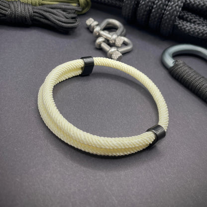 Cream Milan rope bracelet with black sliding clasps, displayed on a dark grey surface with EDC gear in the background.