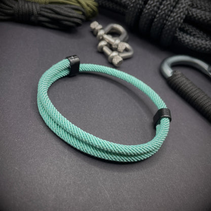 Mint Green Milan rope bracelet with black sliding clasps, displayed on a dark grey surface with EDC gear in the background.