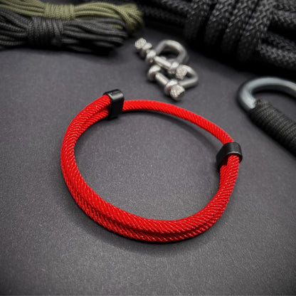 Red Milan rope bracelet with black sliding clasps, displayed on a dark grey surface with EDC gear in the background.