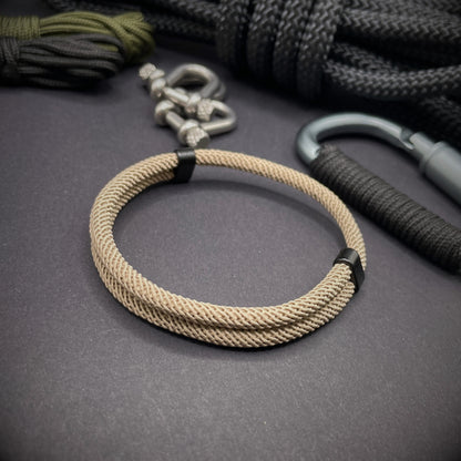 Tan Milan rope bracelet with black sliding clasps, displayed on a dark grey surface with EDC gear in the background.