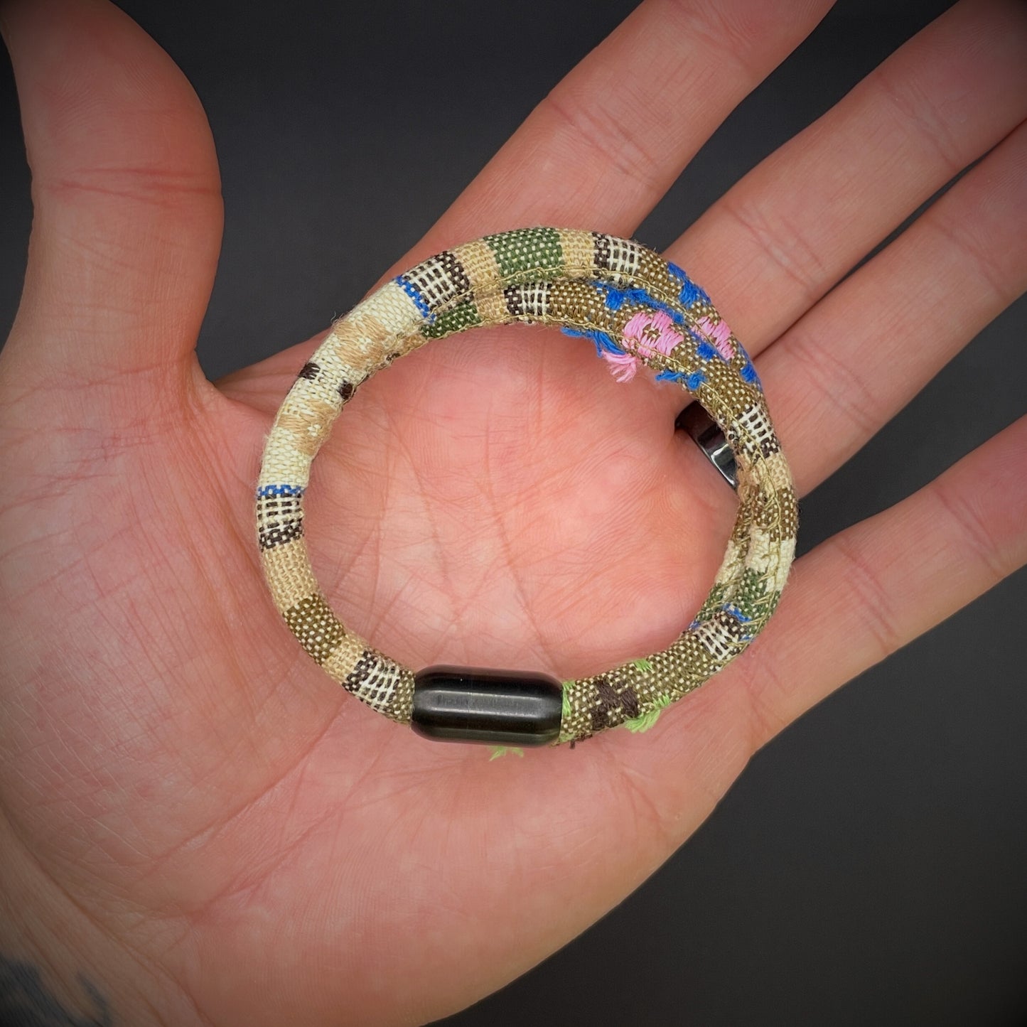 Khaki Boho rope bracelet with black magnetic clasps, displayed in an open palmed hand over a dark grey surface.