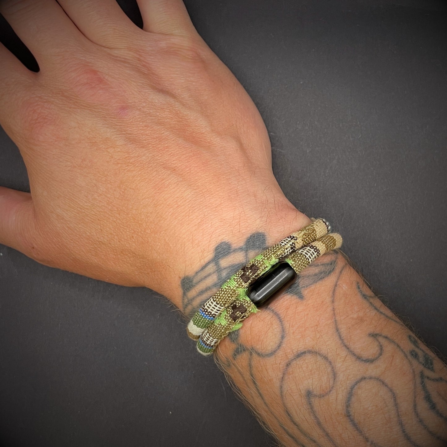 Khaki Boho rope bracelet with black magnetic clasps, displayed on a tattooed wrist over a dark grey surface.