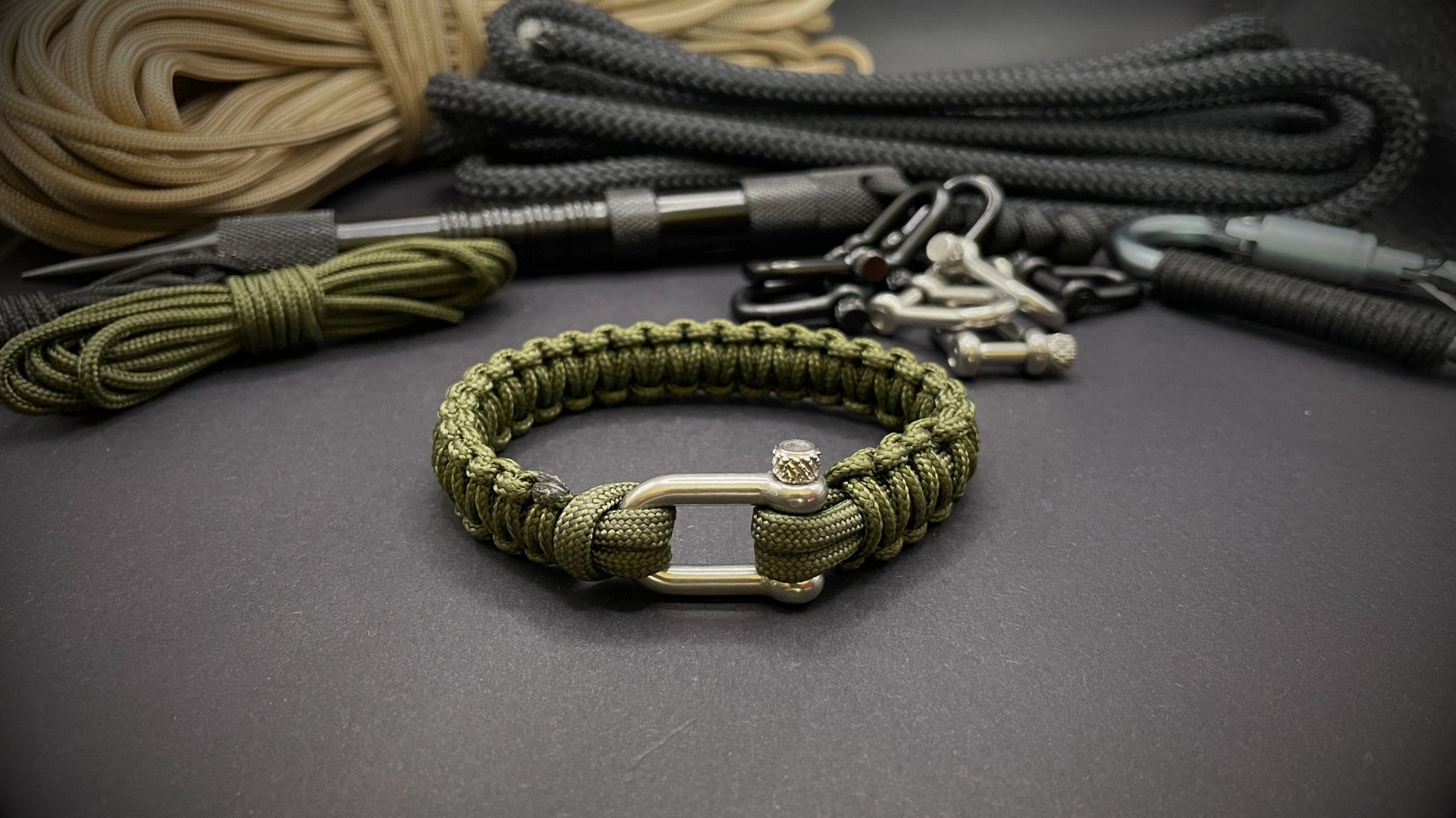 Army green 275 paracord bracelet with stainless steel shackle clasp, displayed on a dark grey surface with EDC gear in the background.