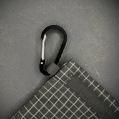 Black aluminium carabiner attached to a black grid-patterned pouch, arranged on a dark surface.