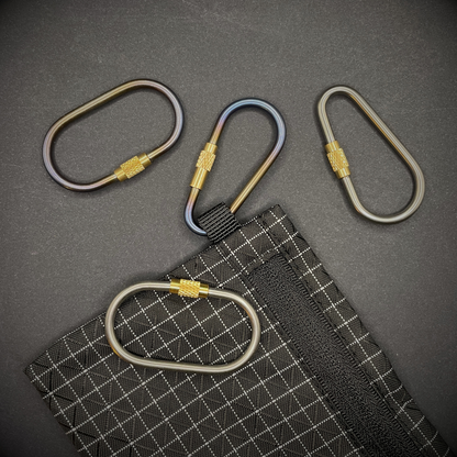 A selection of Mini Locking Titanium Carabiner, one is attached to a black grid-patterned pouch, arranged on a dark surface.