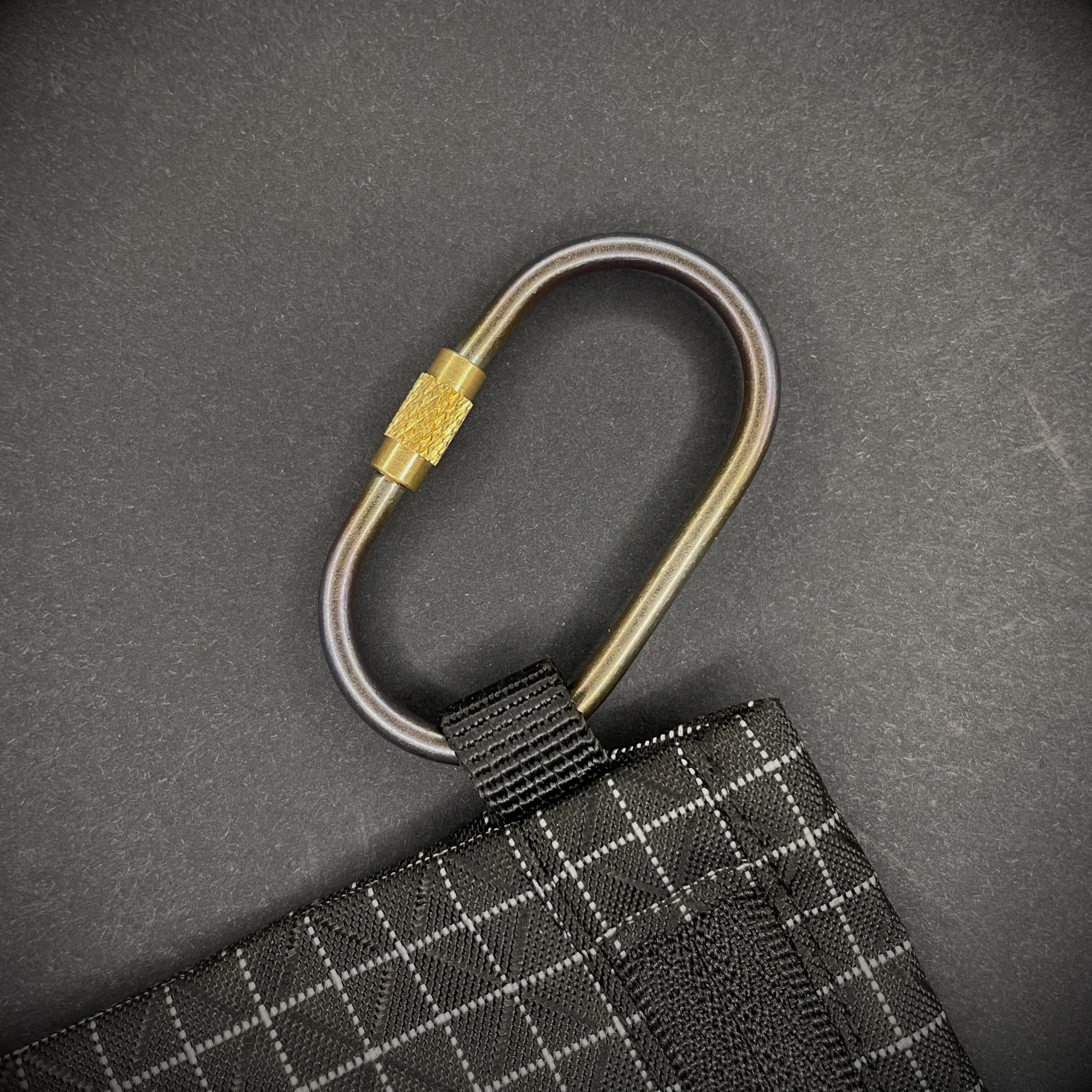 Mini Locking Titanium Carabiner attached to a black grid-patterned pouch, arranged on a dark surface.