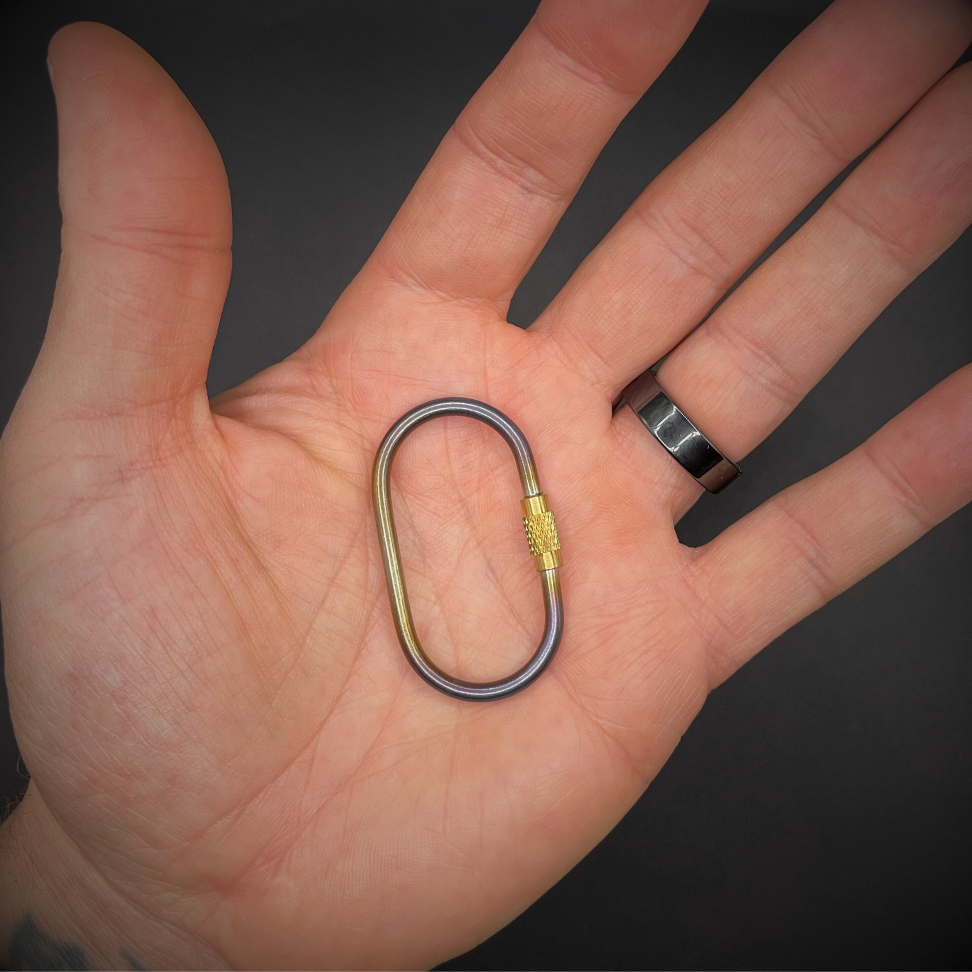 Mini Locking Titanium Carabiner, one is held in the centre of an open hand, arranged on a dark surface.