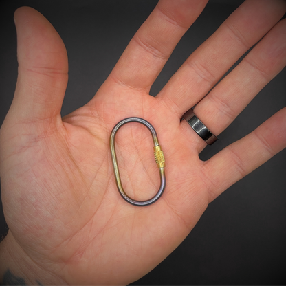Mini Locking Titanium Carabiner, one is held in the centre of an open hand, arranged on a dark surface.