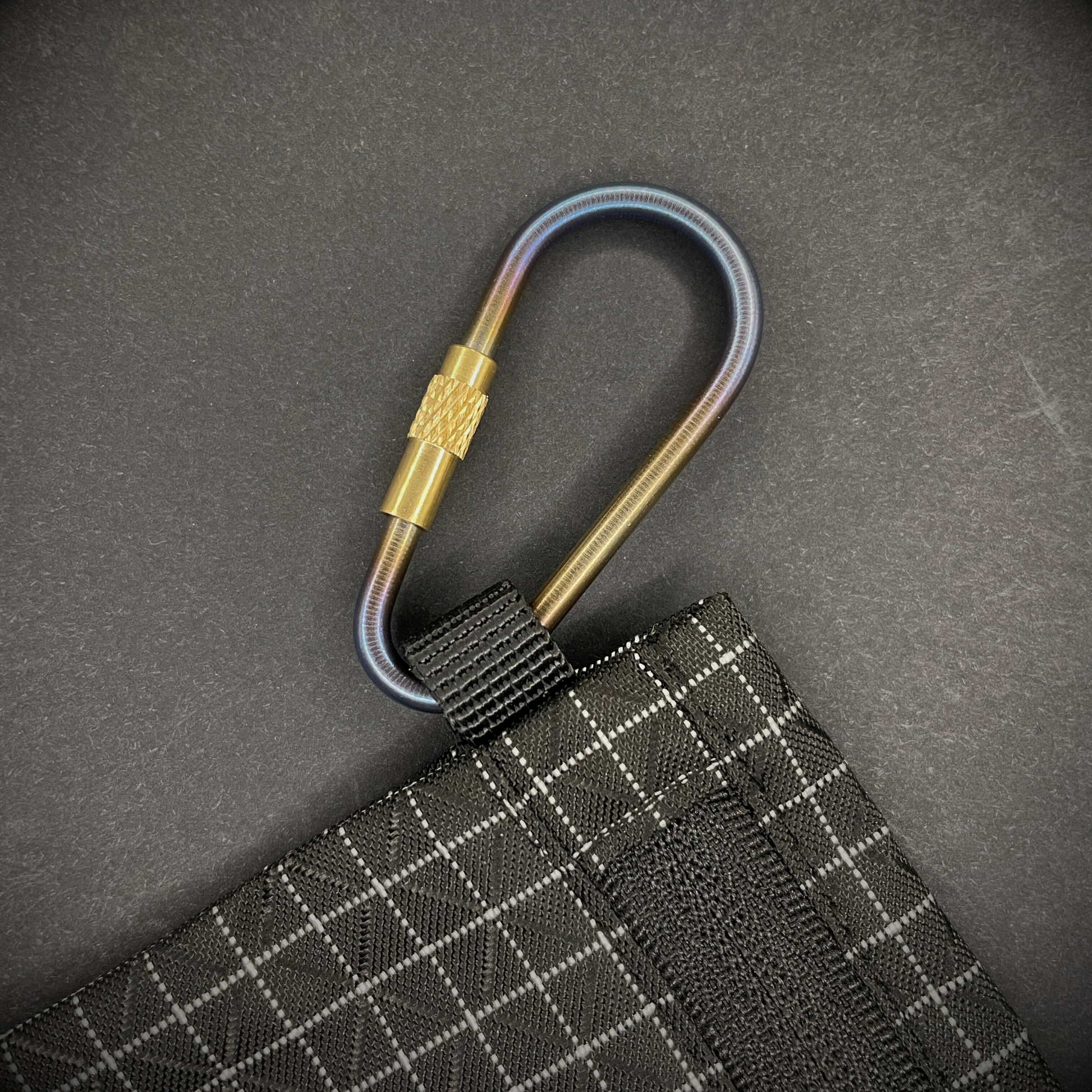 Mini Locking Titanium Carabiner attached to a black grid-patterned pouch, arranged on a dark surface.