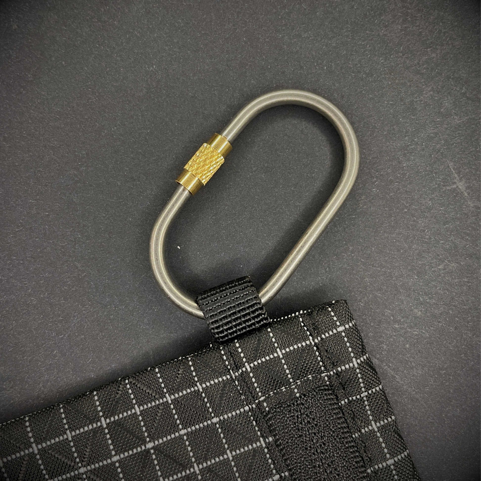 Mini Locking Titanium Carabiner attached to a black grid-patterned pouch, arranged on a dark surface.
