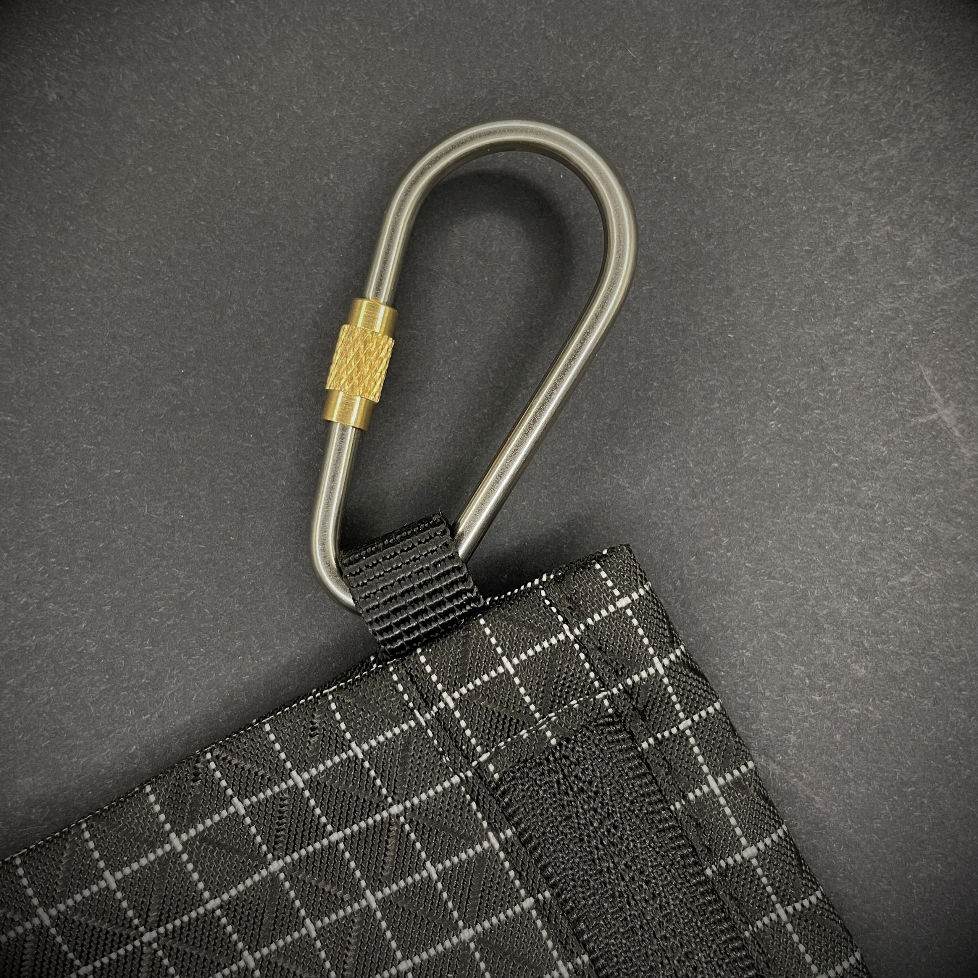 Mini Locking Titanium Carabiner attached to a black grid-patterned pouch, arranged on a dark surface.