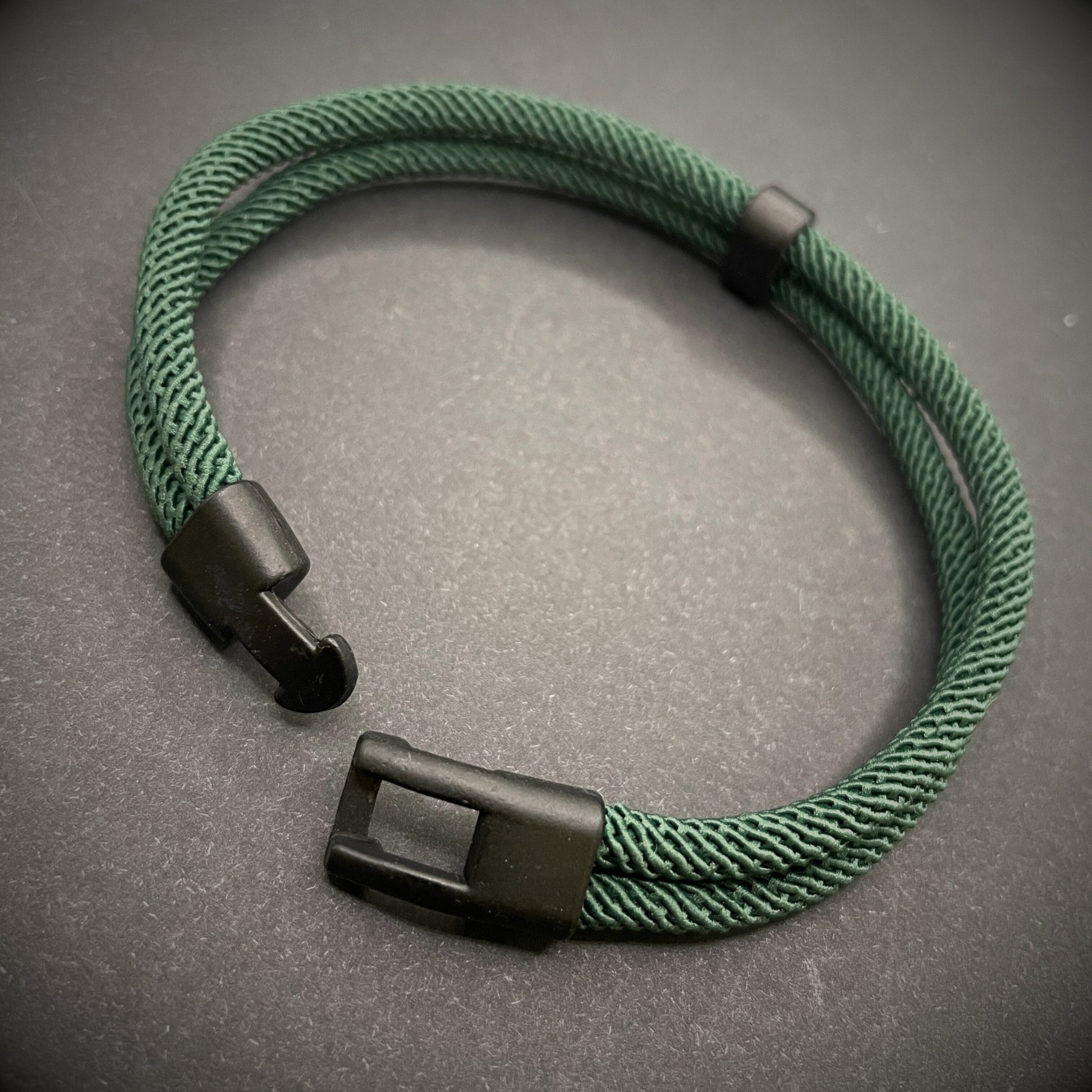 Green rope bracelet with a black metal side-release buckle and minimalist loop detail, displayed on a dark background.