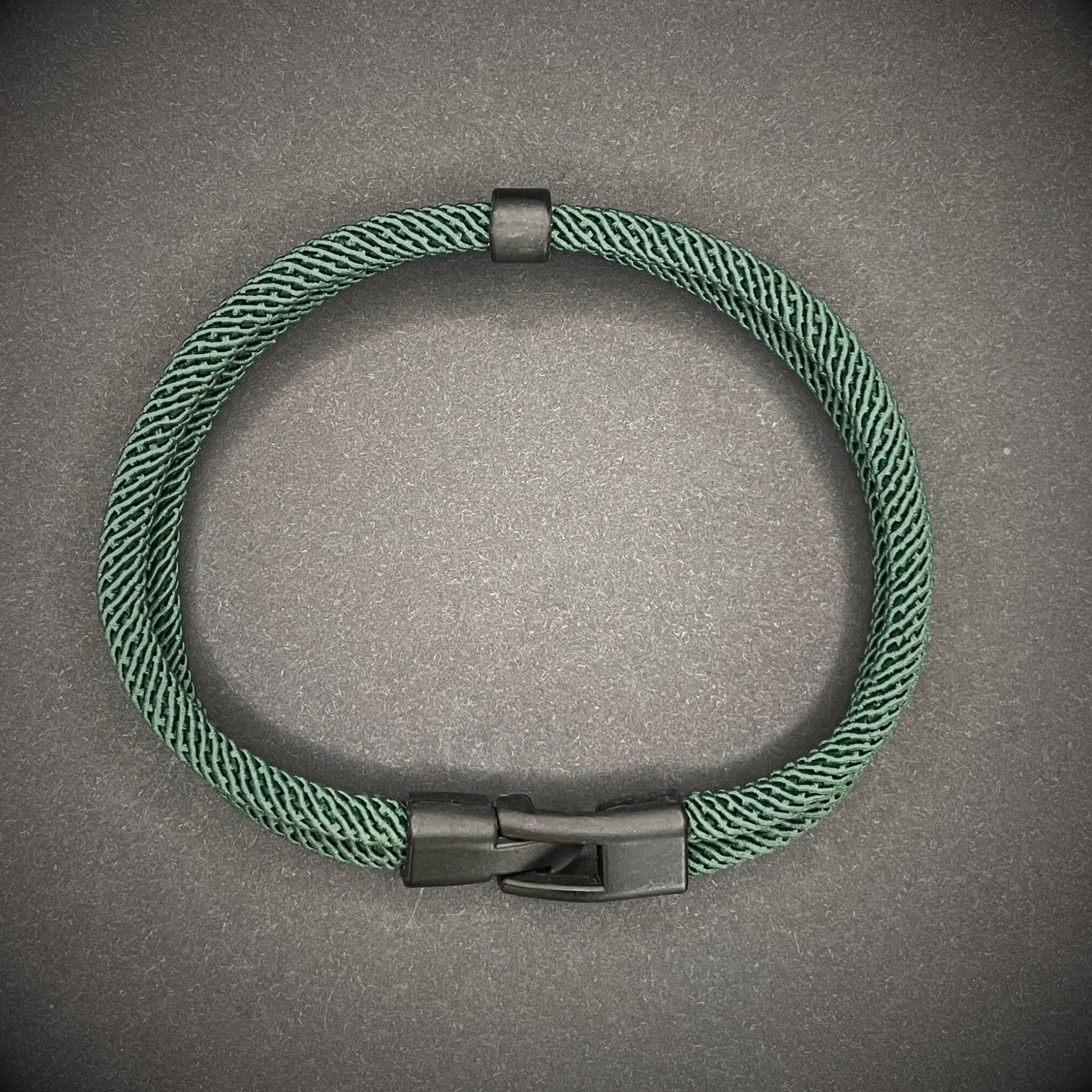 Green rope bracelet with a black metal side-release buckle and minimalist loop detail, displayed on a dark background.