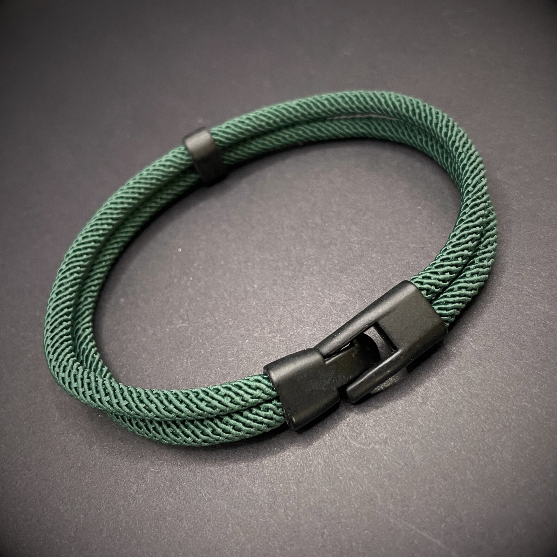Green rope bracelet with a black metal side-release buckle and minimalist loop detail, displayed on a dark background.