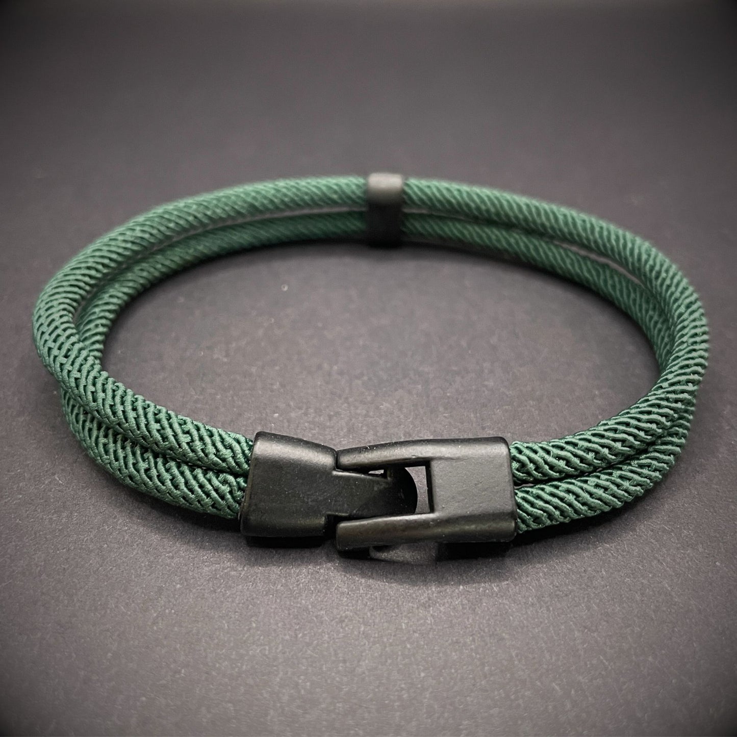 Green rope bracelet with a black metal side-release buckle and minimalist loop detail, displayed on a dark background.