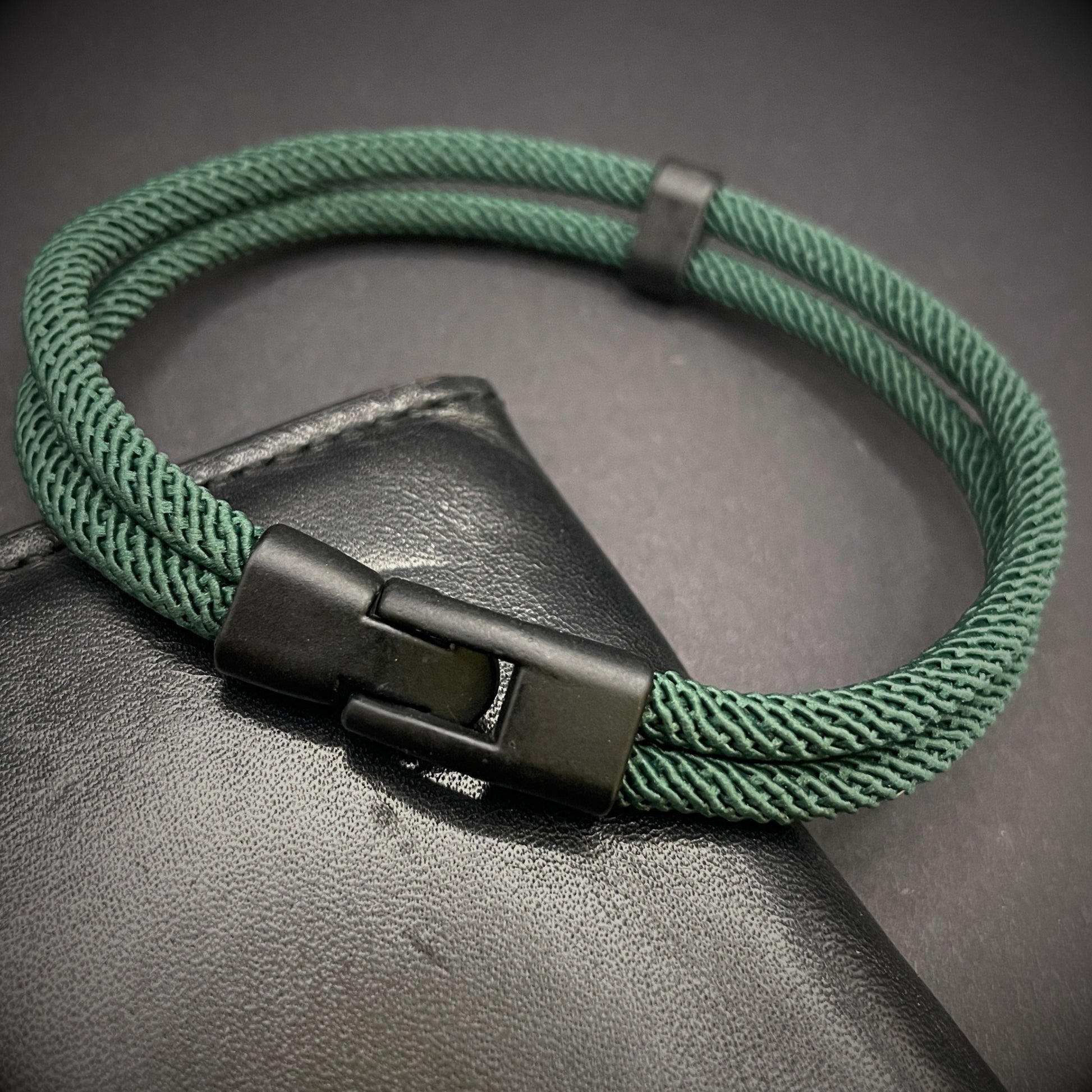 Green rope bracelet with a black metal side-release buckle and minimalist loop detail, displayed on a dark background.
