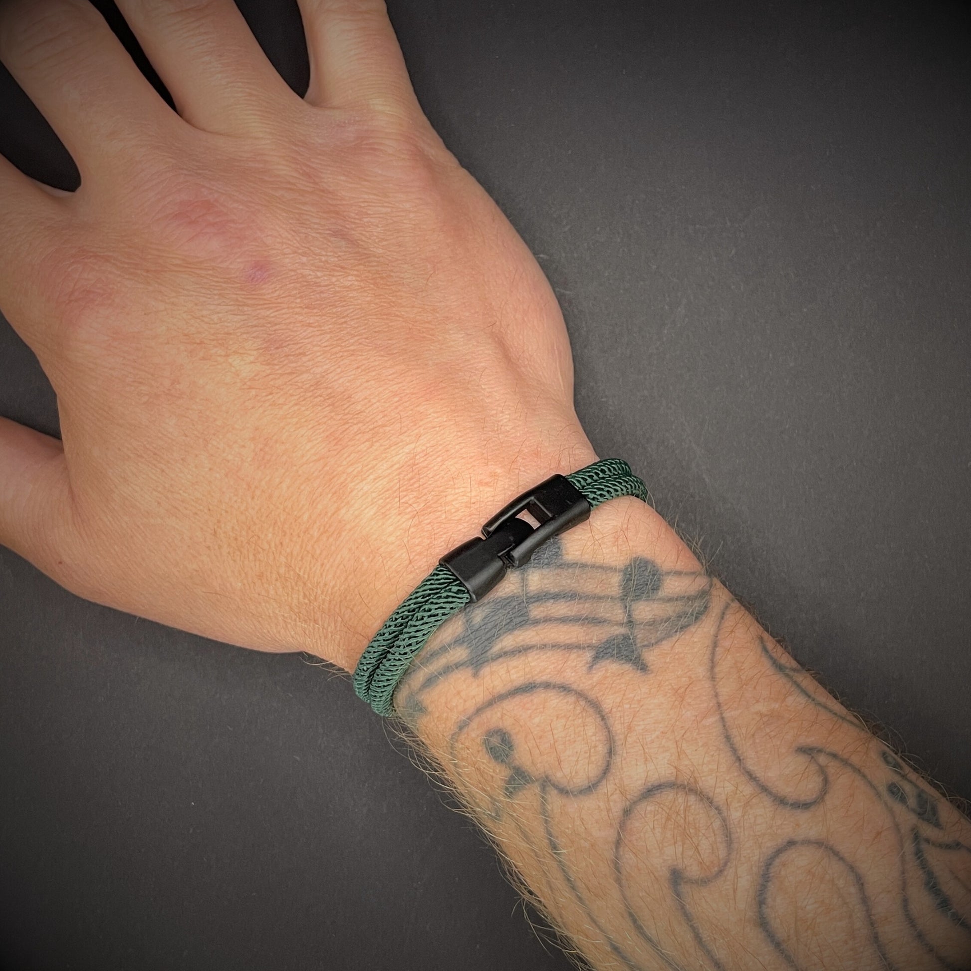 Green rope bracelet with a black metal side-release buckle and minimalist loop detail, displayed on a dark background.