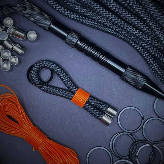 Flat lay of black braided rope keychain with silver end caps and orange whipping, surrounded by paracord supplies including a black metal tool, orange cord, metal rings, and cord ends on a dark background.