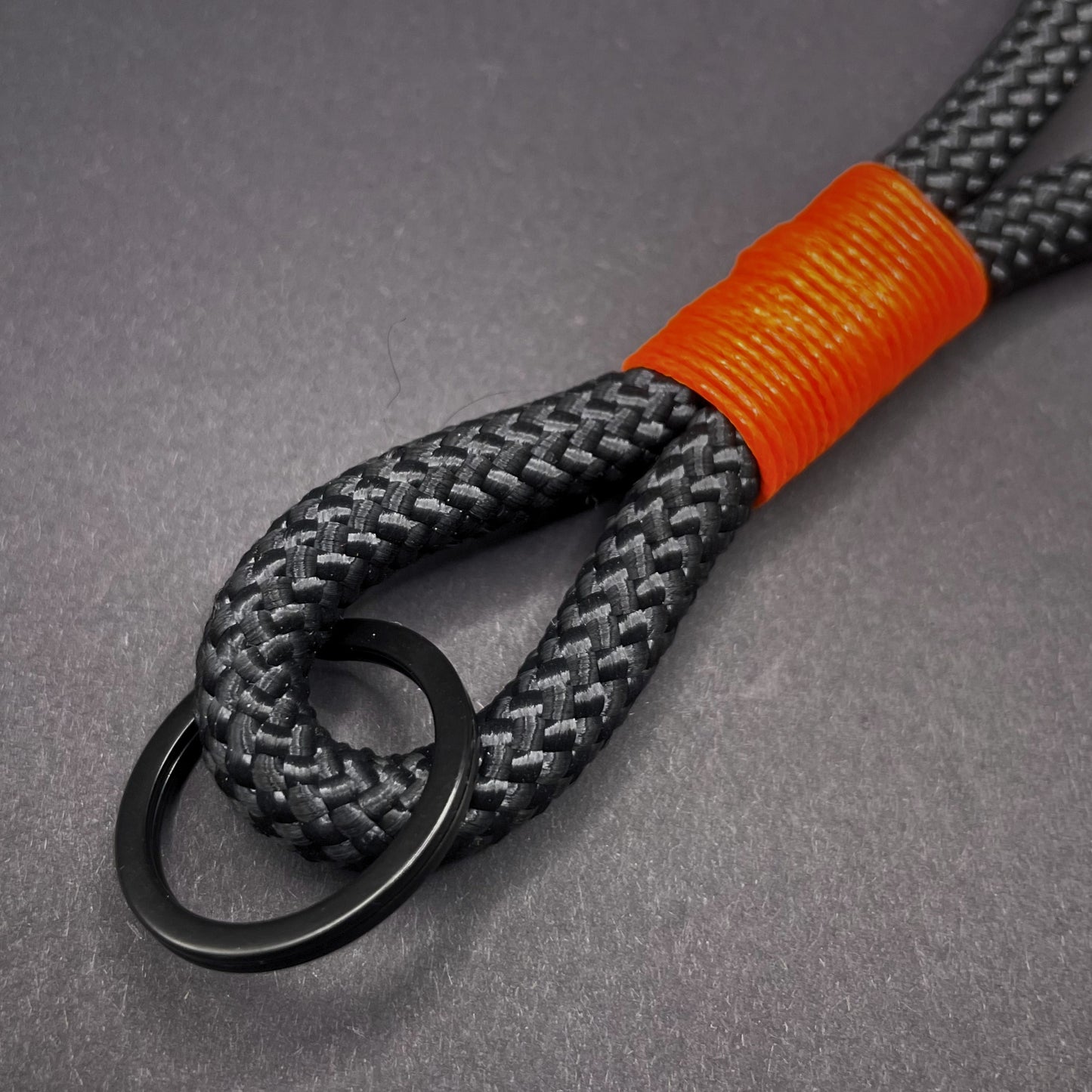 Close up of a black braided rope lanyard with a orange whipping detail, styled on a dark background.