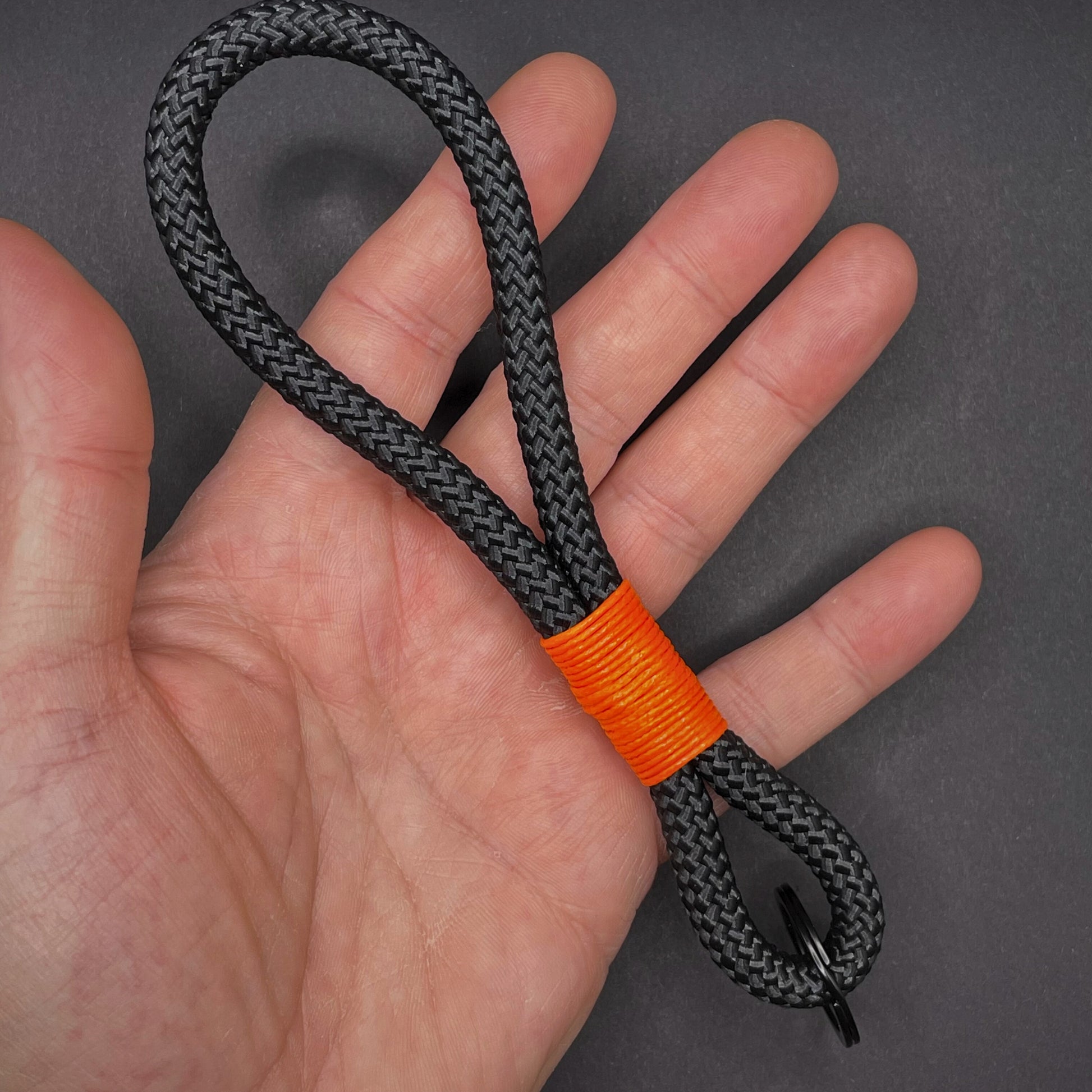Black braided rope lanyard with a orange whipping detail held in a open palmed hand, styled on a dark background.