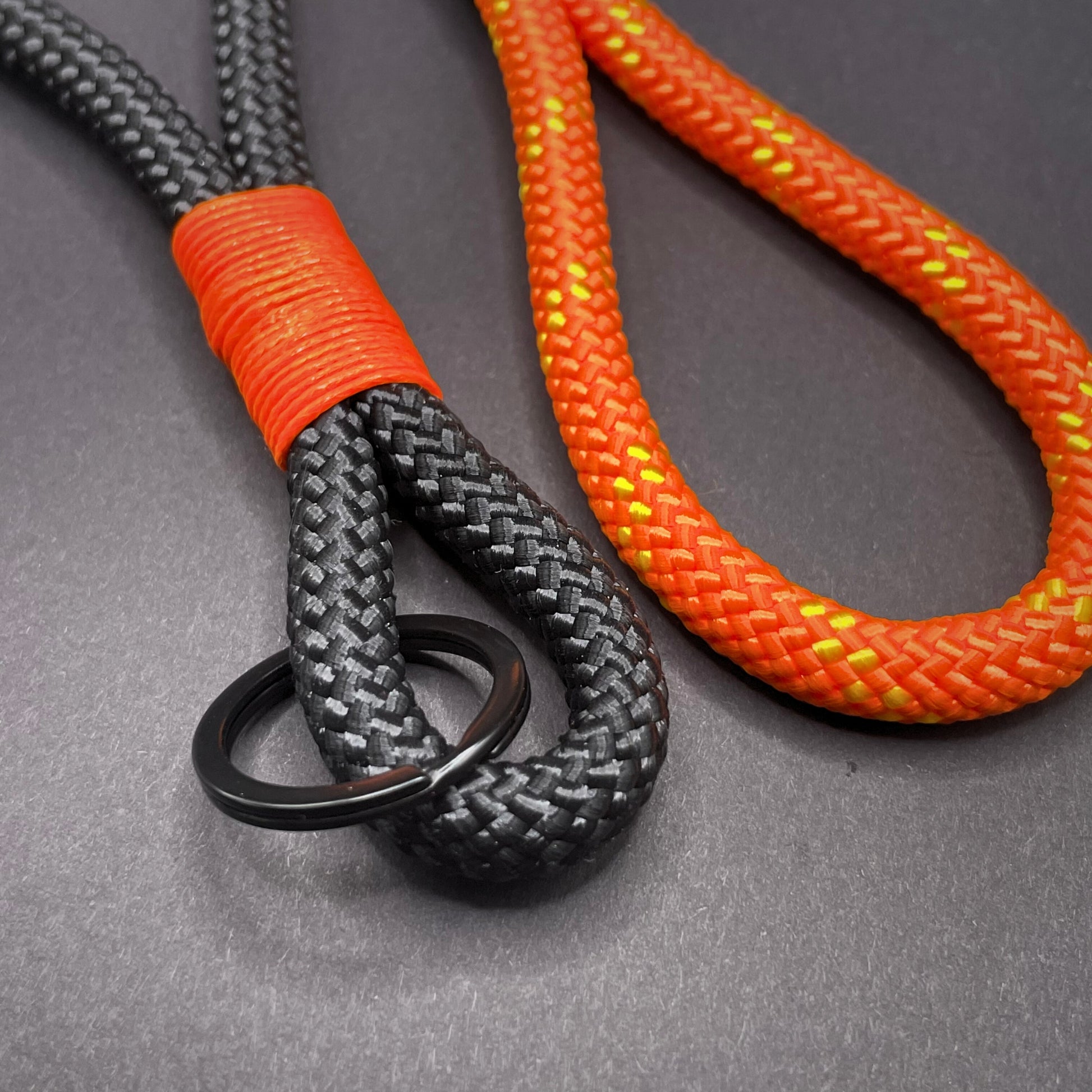 Close up of two braided rope lanyards one with black rope and orange whipping detail, the other with orange rope, styled on a dark background.