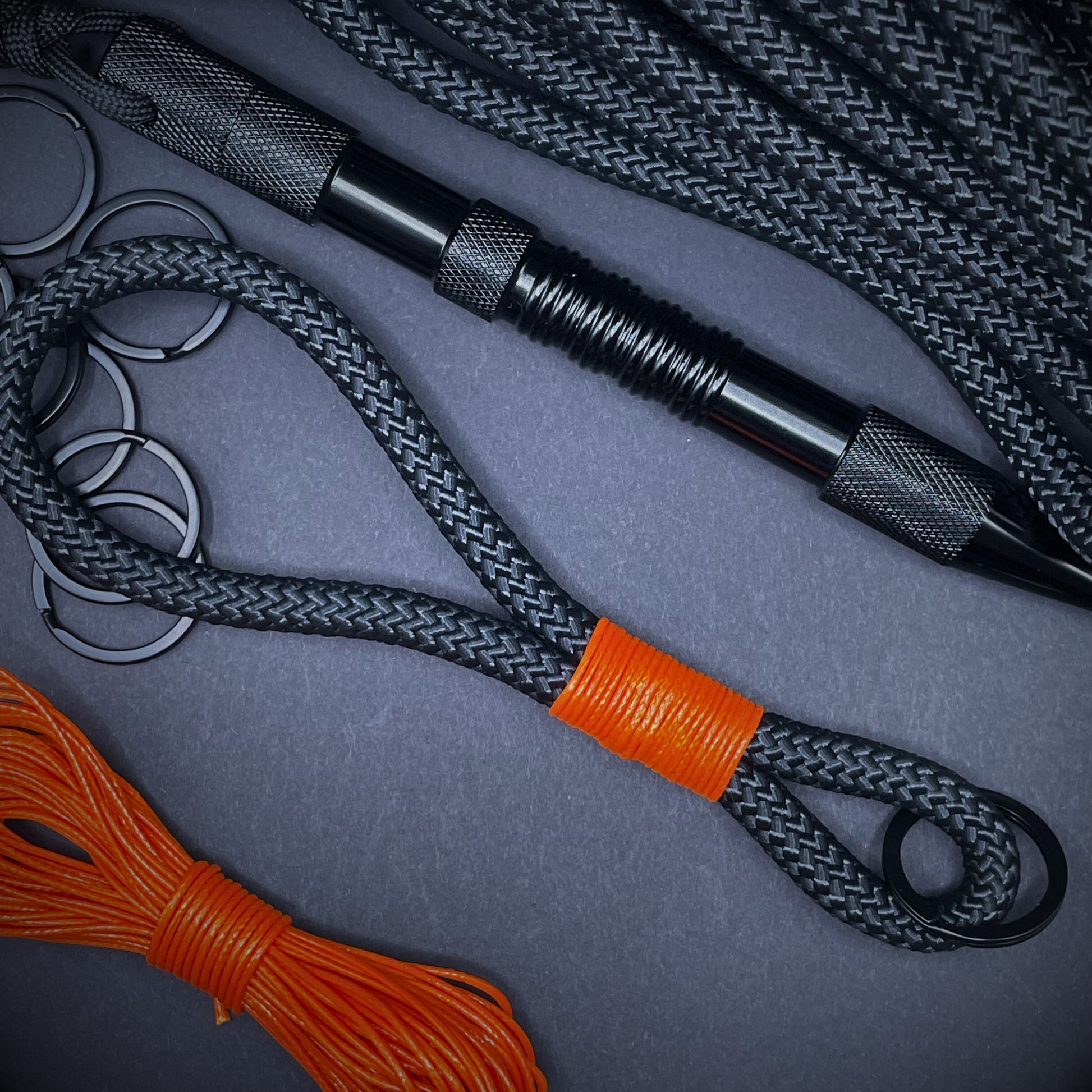 Black braided rope lanyard with a orange whipping detail, styled on a dark background with EDC gear arranged in backdrop.