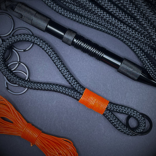 Black braided rope lanyard with a orange whipping detail, styled on a dark background with EDC gear arranged in backdrop.