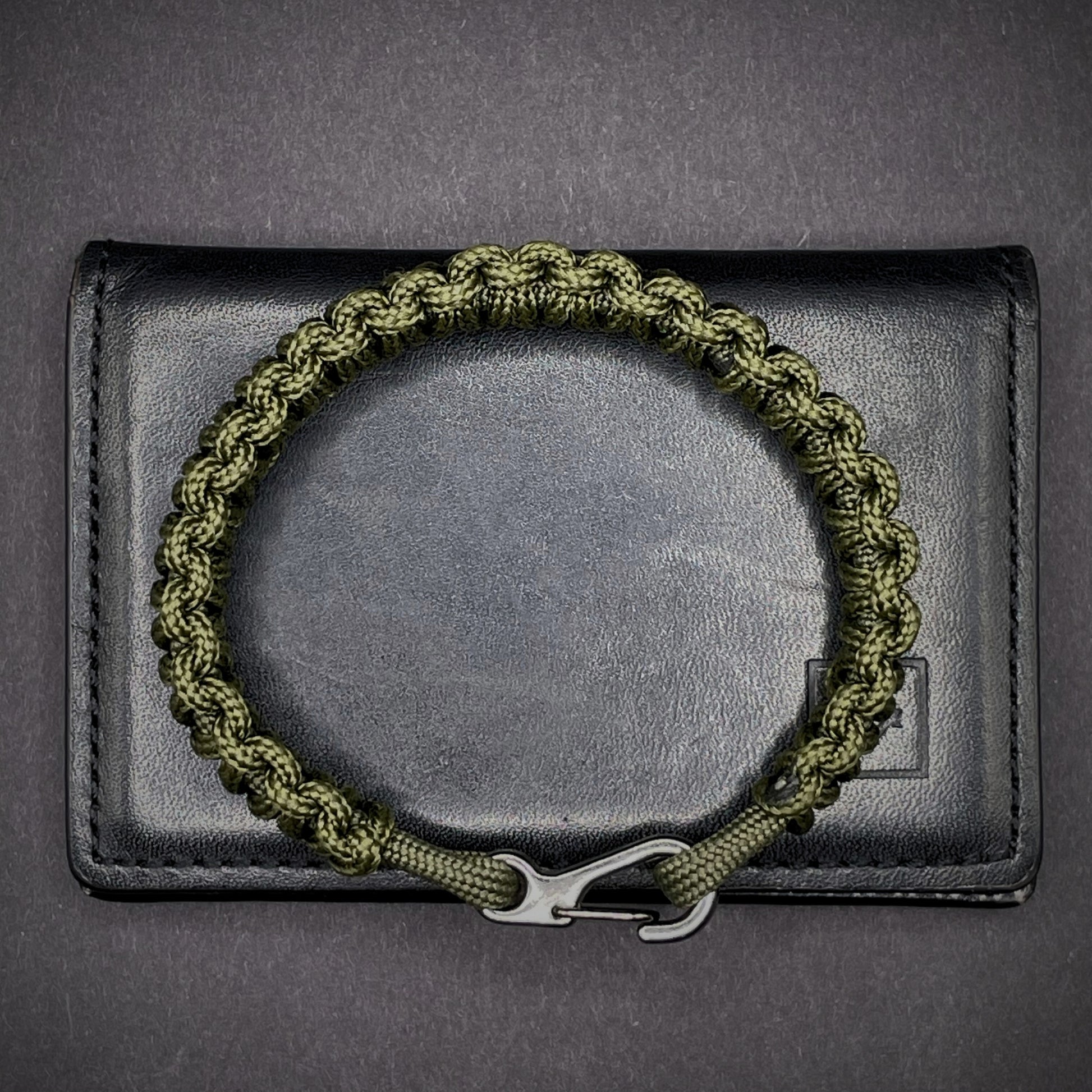 Flat lay of Army Green cobra knot paracord bracelet on a dark grey surface surrounded by EDC items