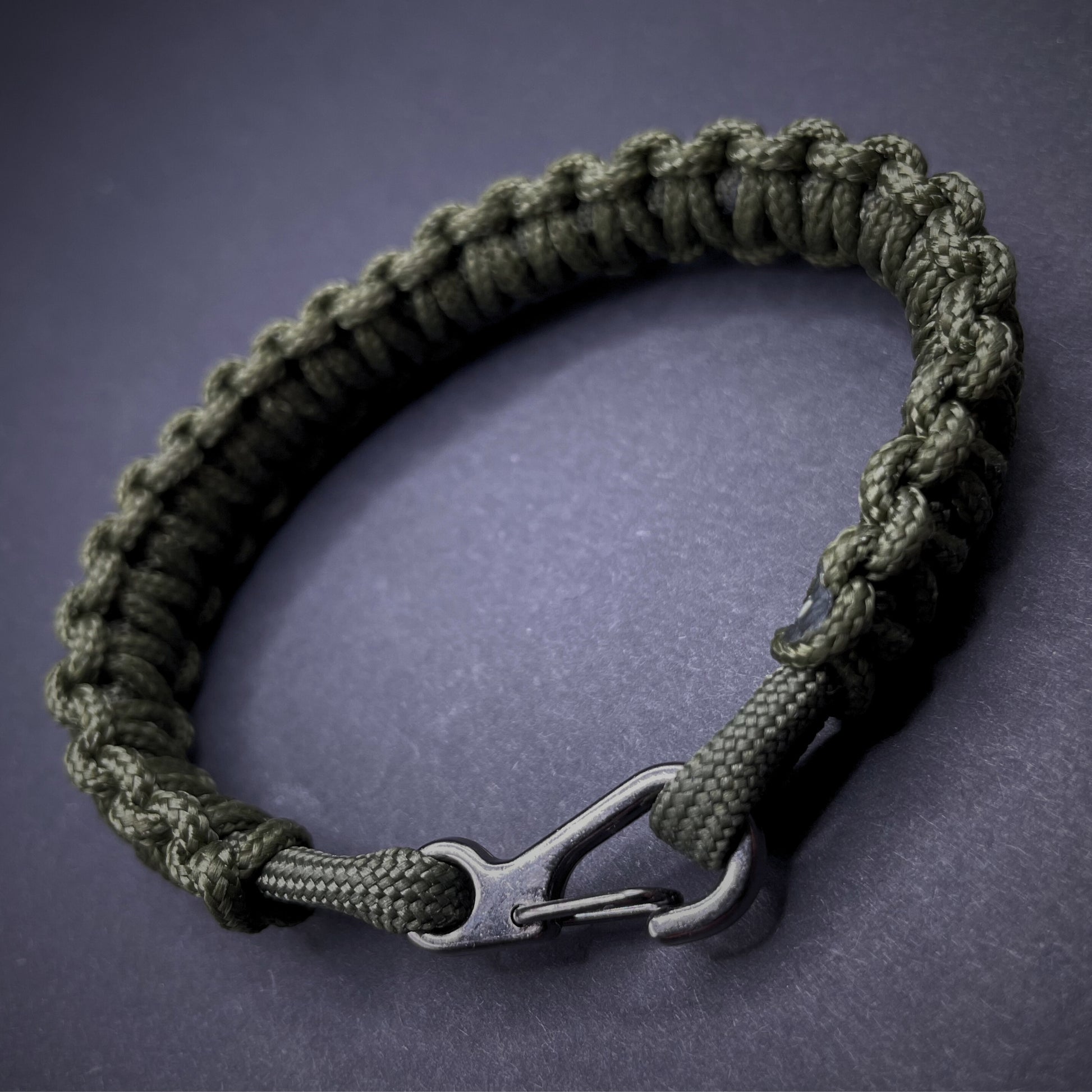 Flat lay of Army Green cobra knot paracord bracelet on a dark grey surface surrounded by EDC items