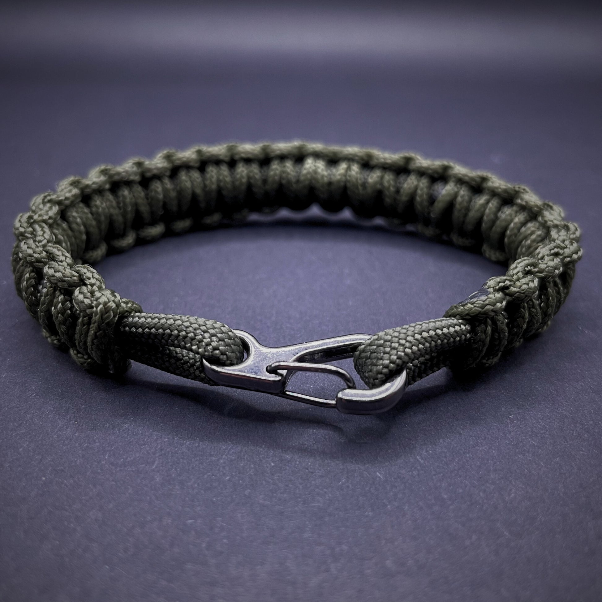 Flat lay of Army Green cobra knot paracord bracelet on a dark grey surface surrounded by EDC items
