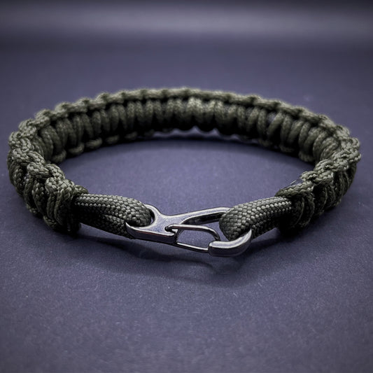 Flat lay of Army Green cobra knot paracord bracelet on a dark grey surface surrounded by EDC items