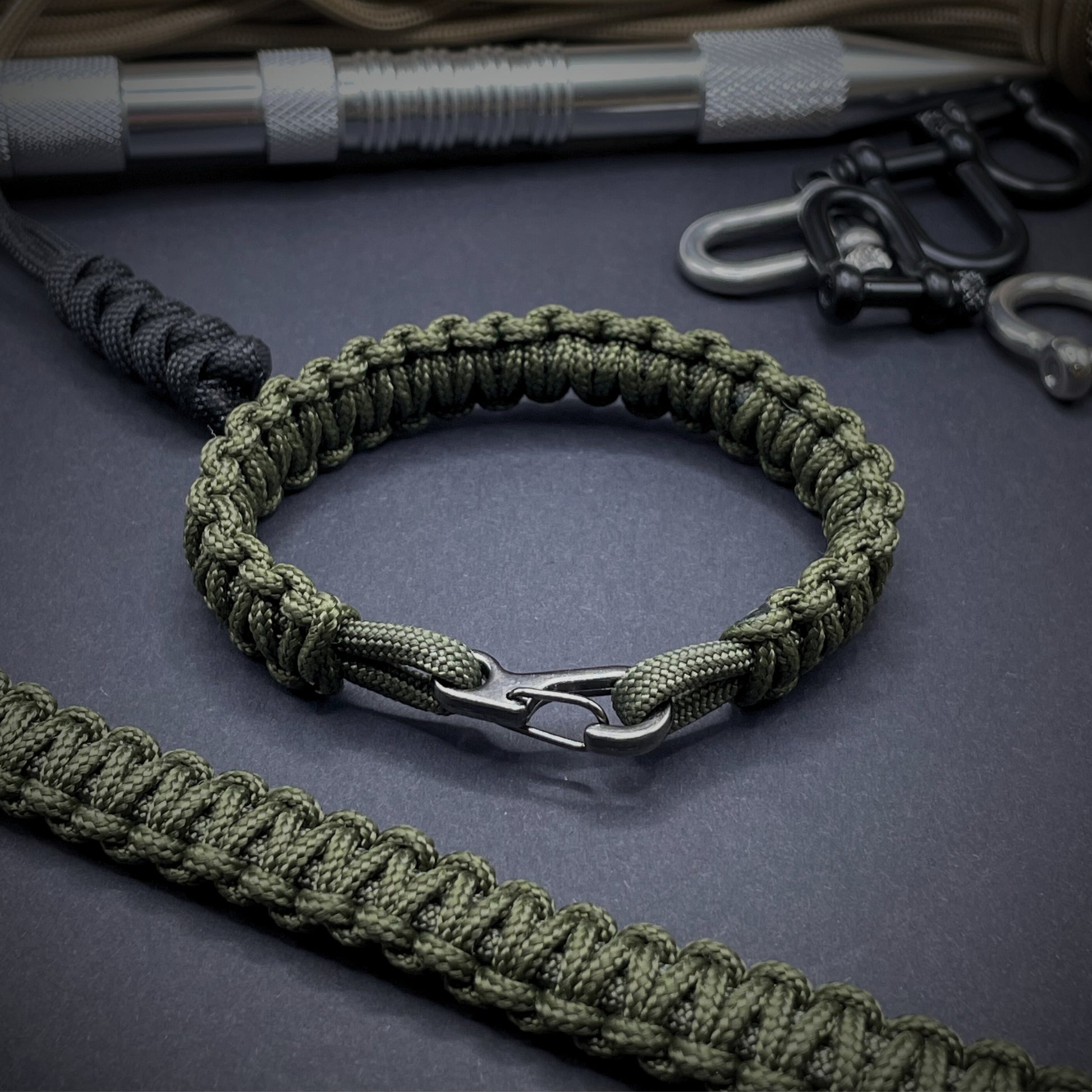 Flat lay of Army Green cobra knot paracord bracelet on a dark grey surface surrounded by EDC items