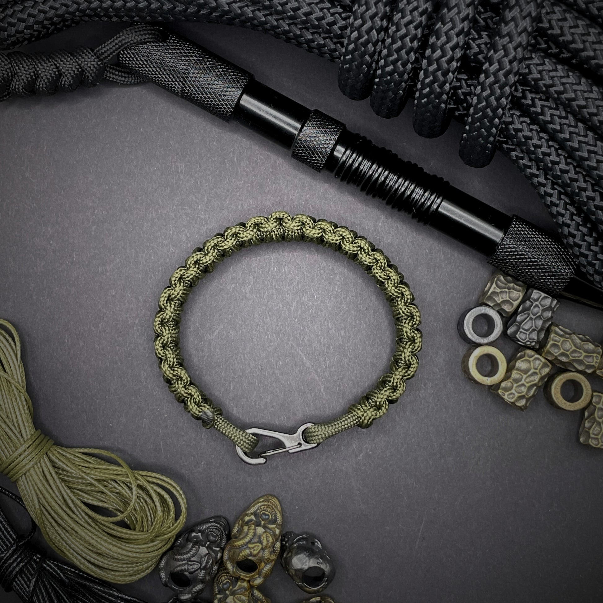 Flat lay of Army Green cobra knot paracord bracelet on a dark grey surface surrounded by EDC items