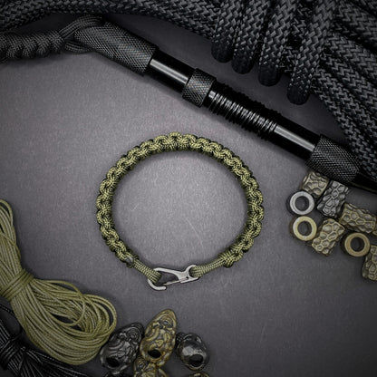 Flat lay of Army Green cobra knot paracord bracelet on a dark grey surface surrounded by EDC items