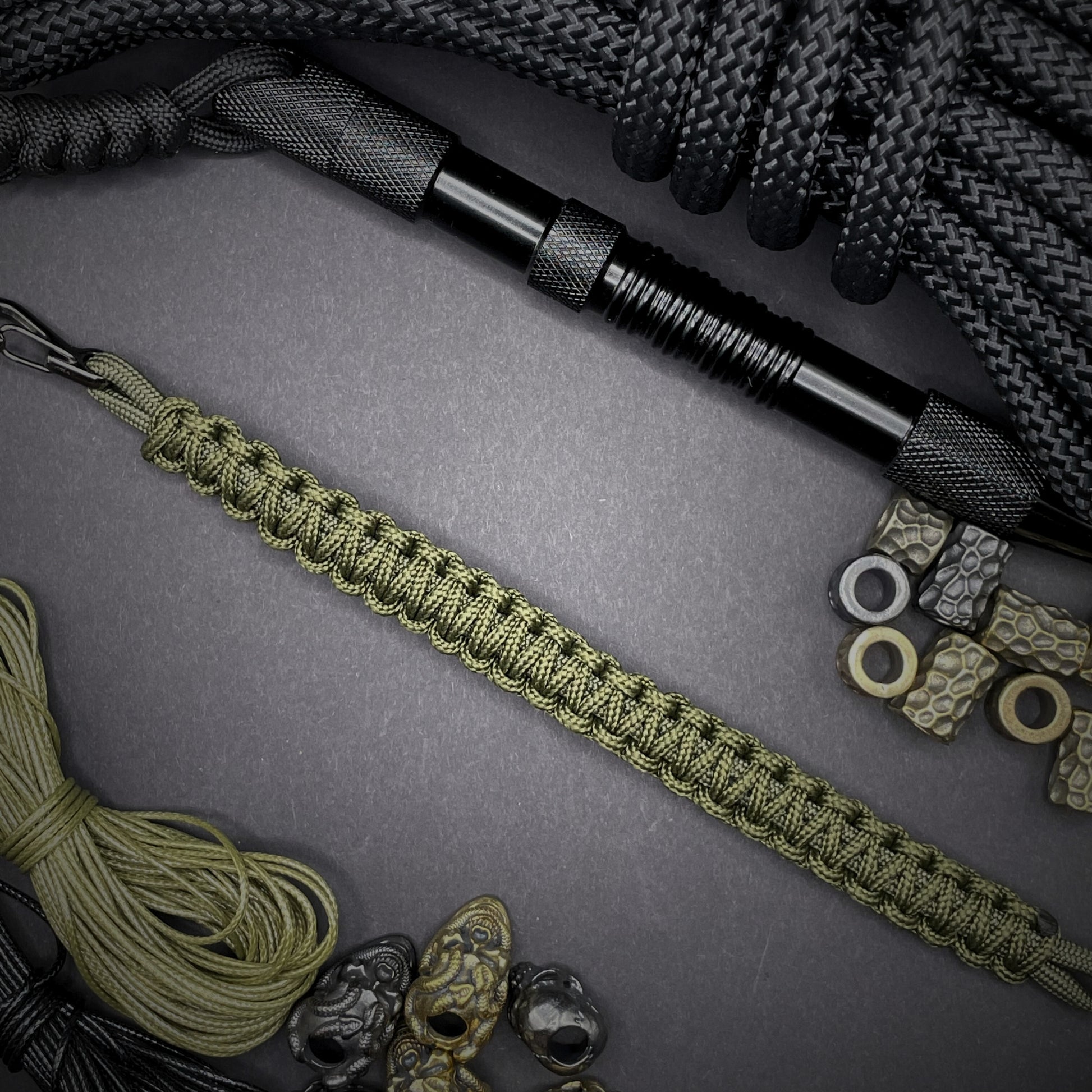 Flat lay of Army Green cobra knot paracord bracelet on a dark grey surface surrounded by EDC items