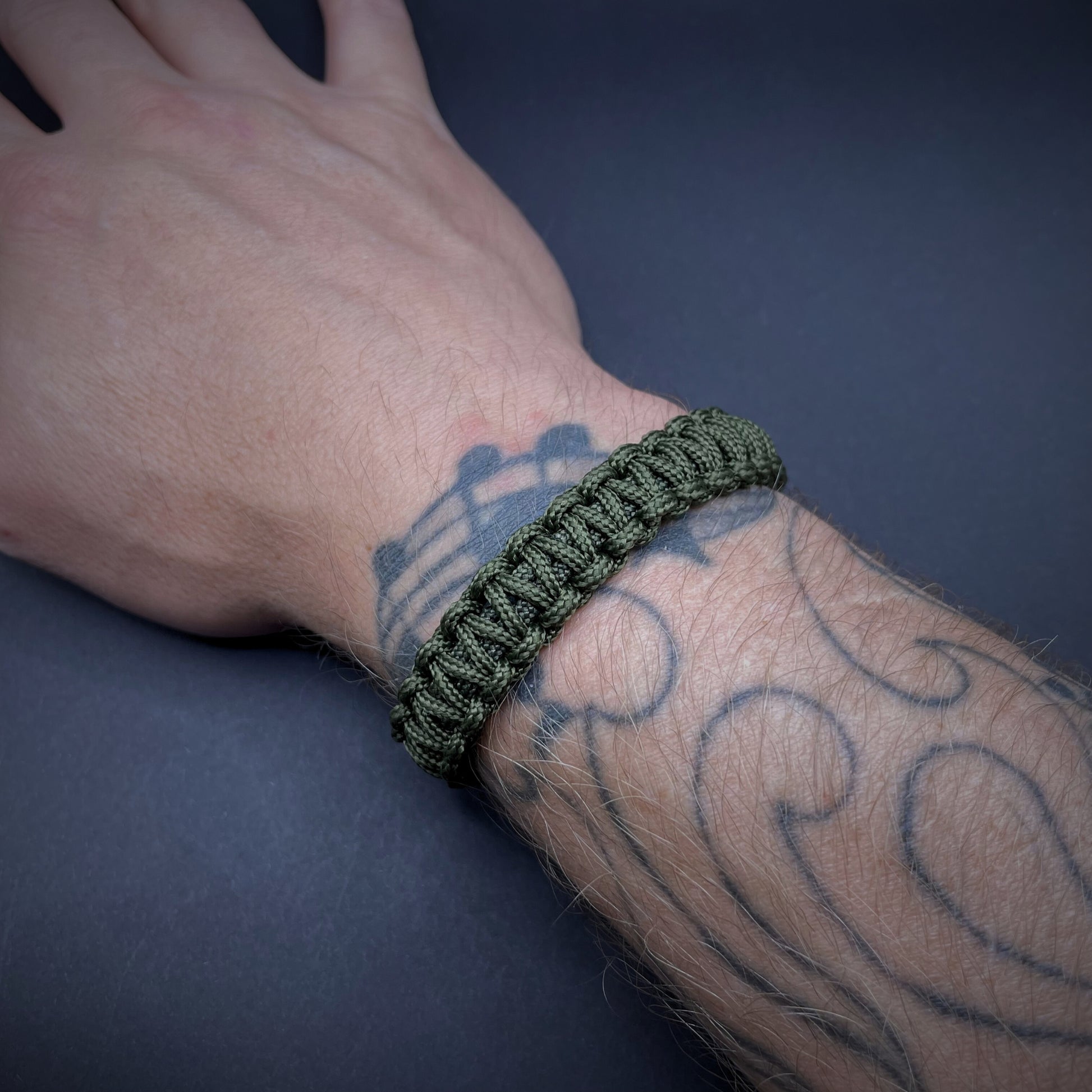Army Green cobra knot paracord bracelet shown on a tattooed wrist over a dark grey surface