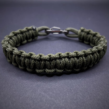 Flat lay of Army Green cobra knot paracord bracelet on a dark grey surface surrounded by EDC items