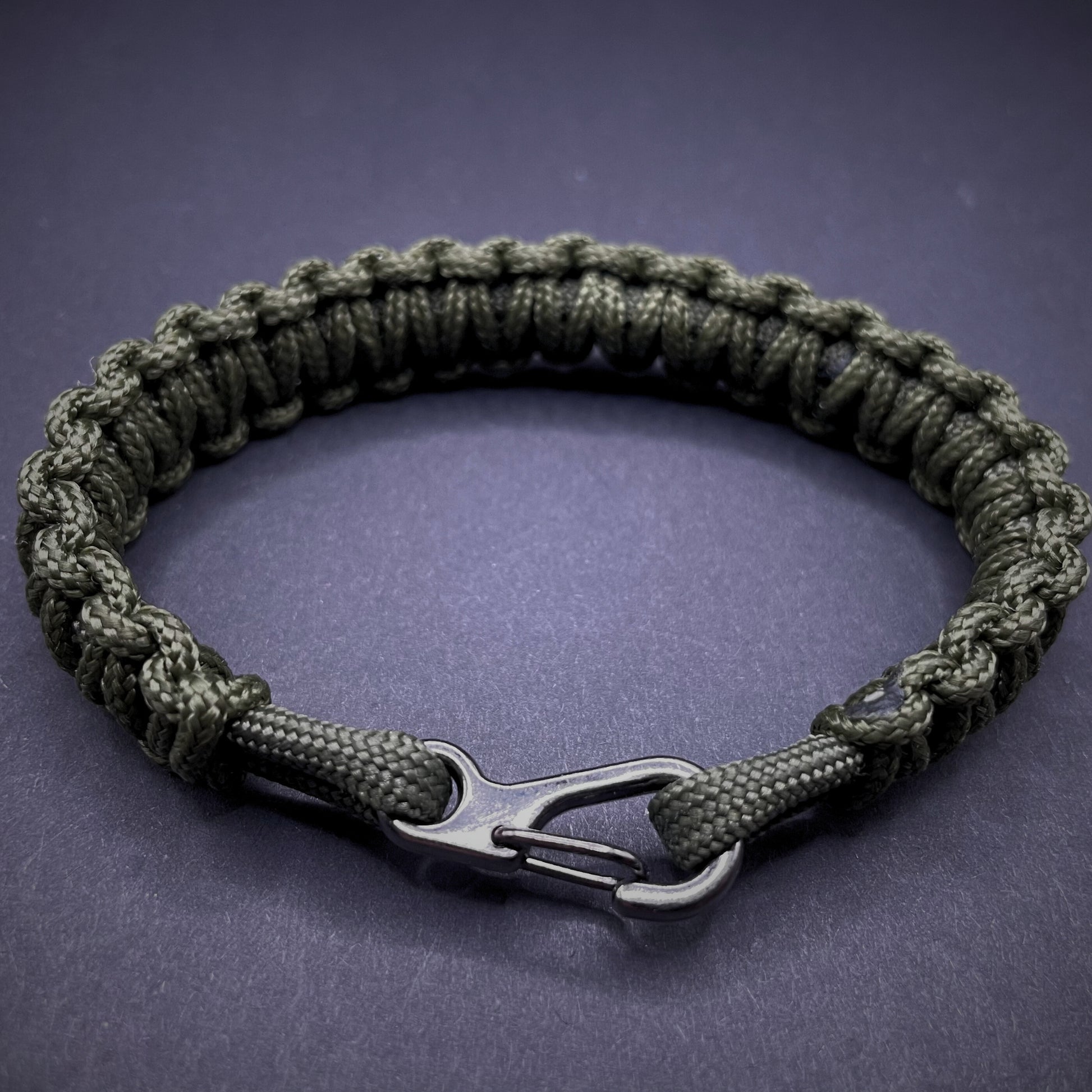 Flat lay of Army Green cobra knot paracord bracelet on a dark grey surface surrounded by EDC items