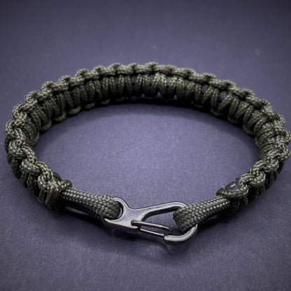 Flat lay of Army Green cobra knot paracord bracelet on a dark grey surface surrounded by EDC items