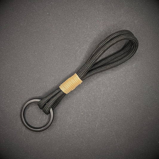 Flat lay of black 550 paracord keychain with tan cotton whipping on a dark background.