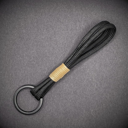Flat lay of black 550 paracord keychain with tan cotton whipping on a dark background.