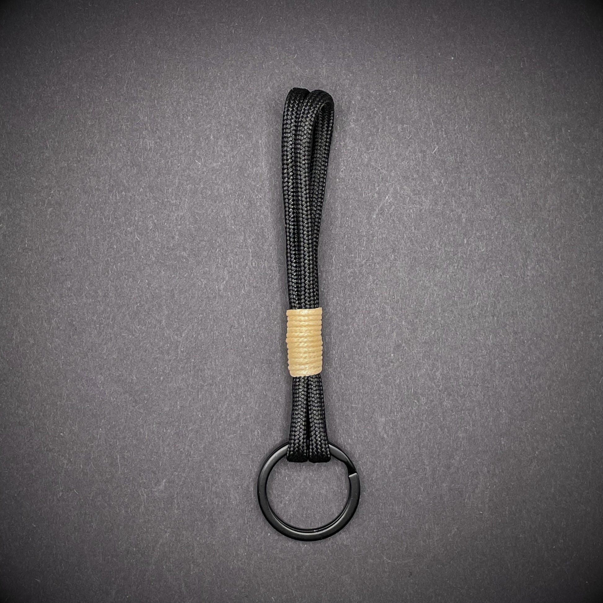 Flat lay of black 550 paracord keychain with tan cotton whipping on a dark background.