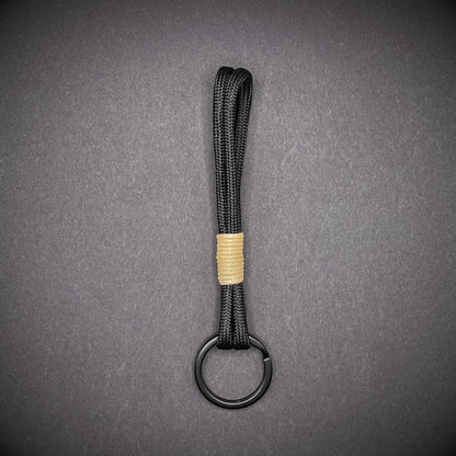 Flat lay of black 550 paracord keychain with tan cotton whipping on a dark background.