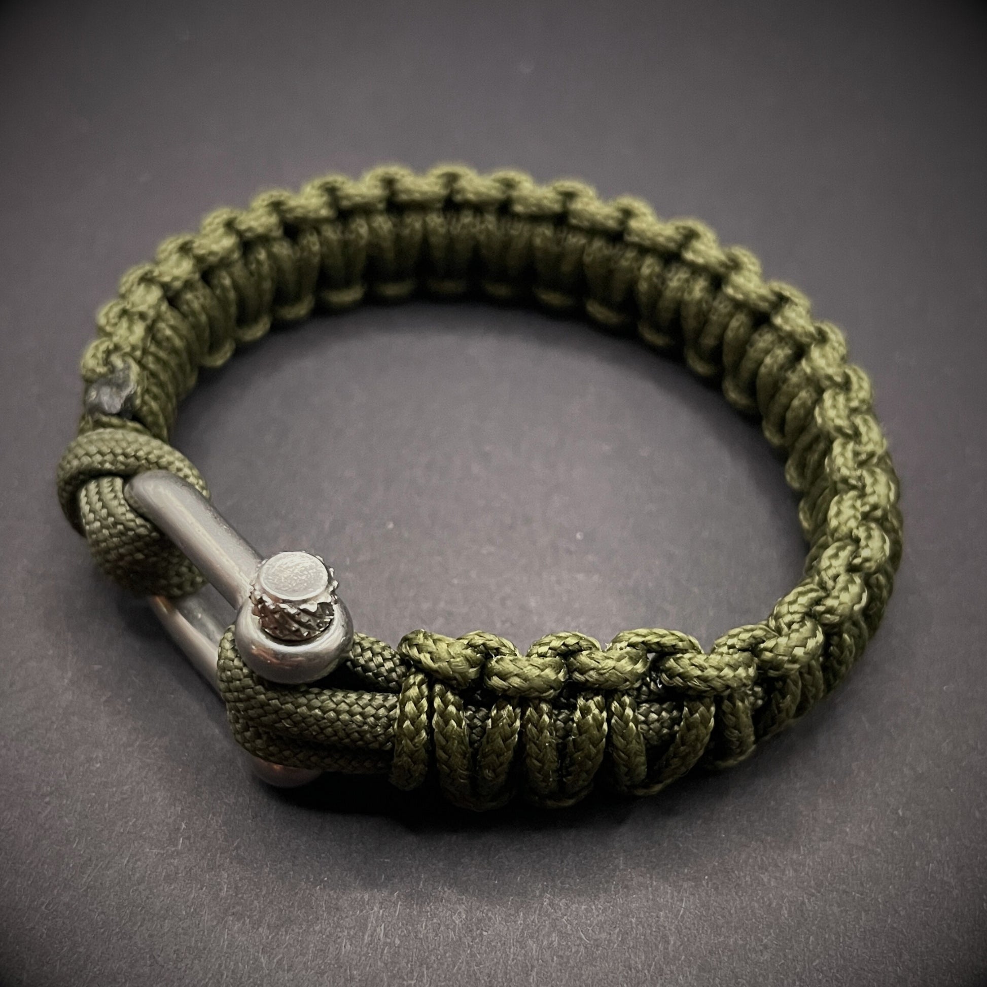An army green paracord rope bracelet arranged on a dark grey surface.