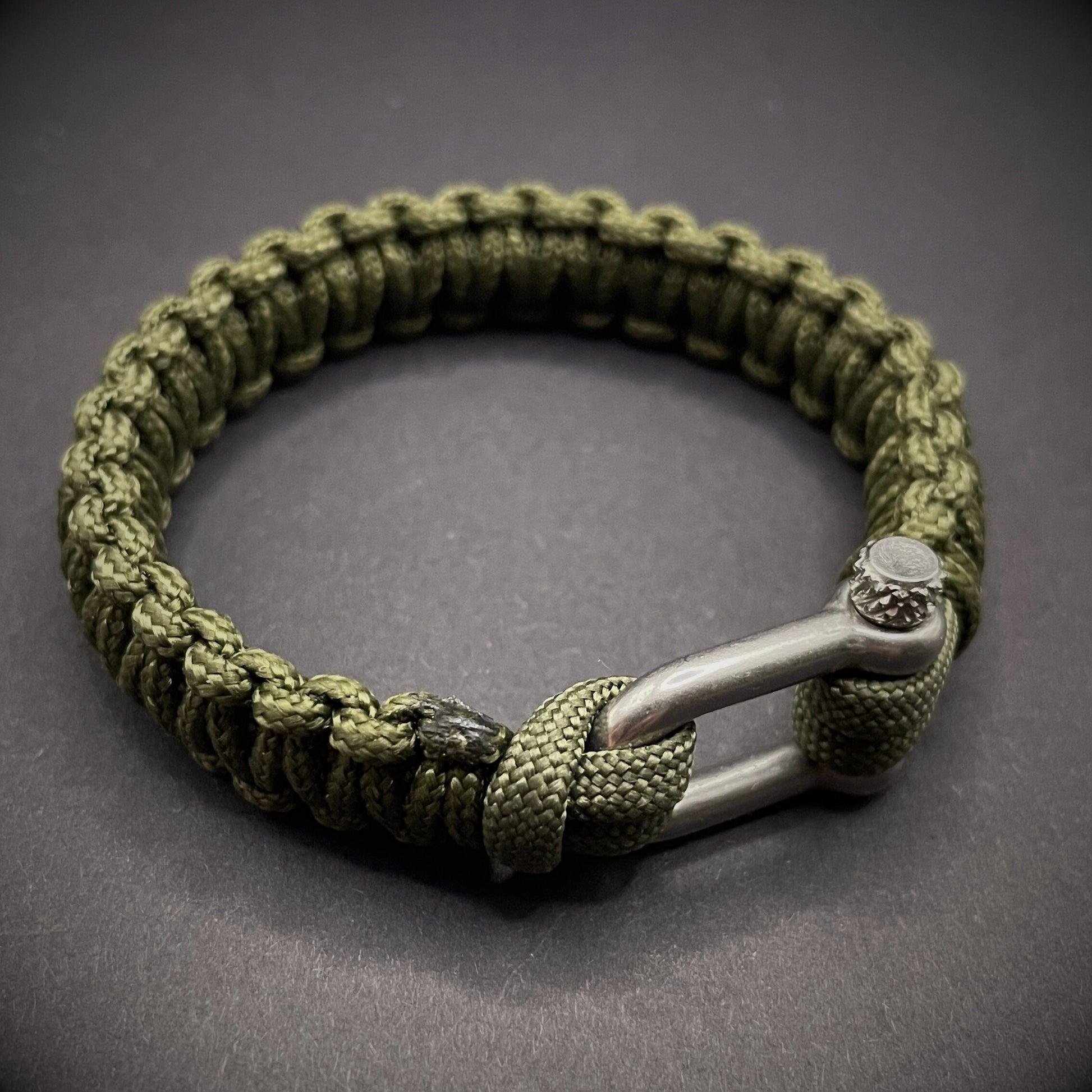 An army green paracord rope bracelet arranged on a dark grey surface.